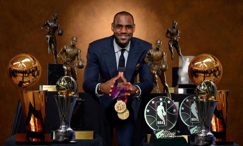 LeBron James – trophées NBA LeBron James posant en costume devant ses trophées : titres de champion NBA, trophées MVP et médailles d’or olympique