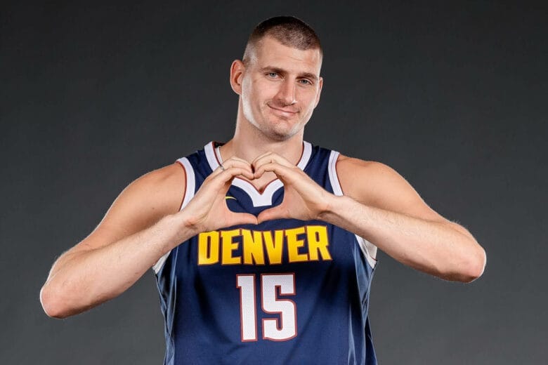 Nikola Jokić forme un cœur avec ses mains en portant le maillot bleu n°15 des Denver Nuggets