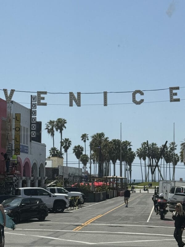 Entrée de Venice Beach avec le célèbre panneau lumineux 'VENICE' suspendu au-dessus de la rue principale, bordée de palmiers