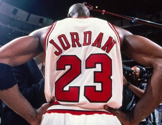 Michael Jordan vu de dos, maillot blanc numéro 23 des Chicago Bulls, bras sur les hanches, sous les projecteurs