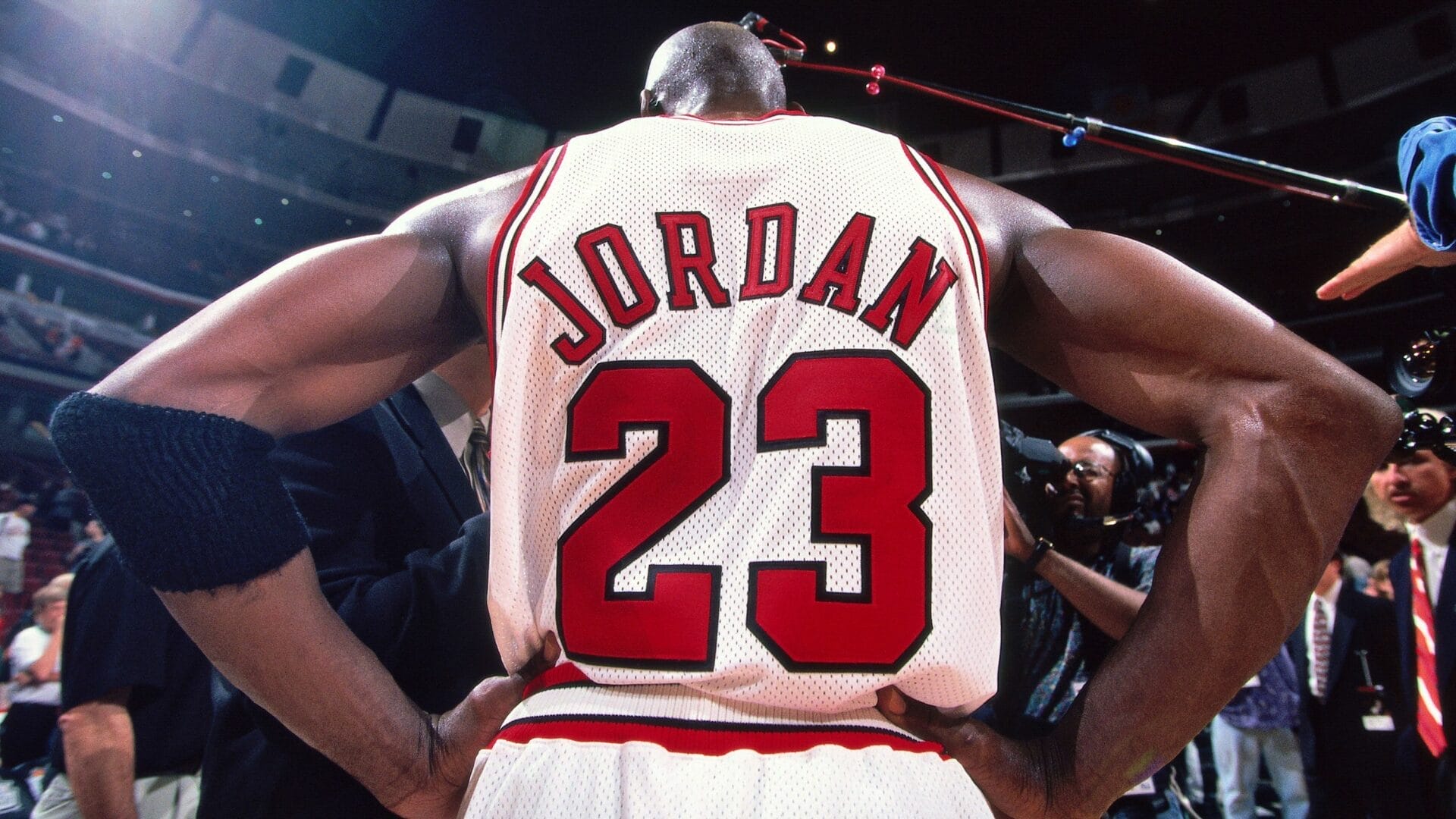Michael Jordan vu de dos, maillot blanc numéro 23 des Chicago Bulls, bras sur les hanches, sous les projecteurs