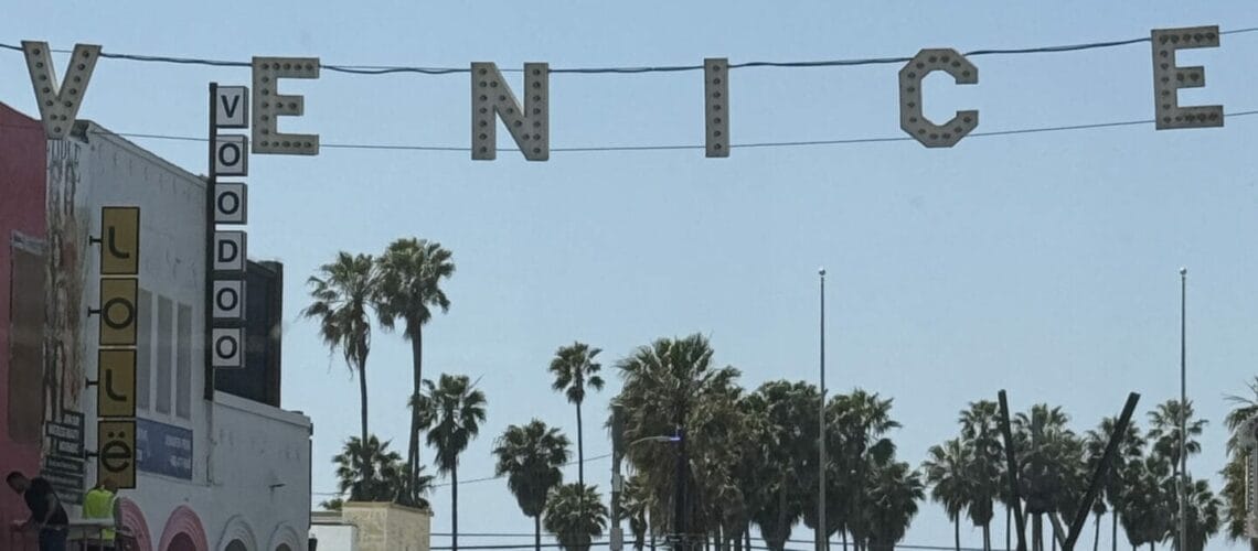 Entrée de Venice Beach avec le célèbre panneau lumineux 'VENICE' suspendu au-dessus de la rue principale, bordée de palmiers